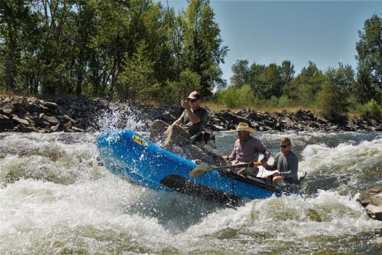 Three people, mid air, on a raft in some rapids. A man on the front is fishing.