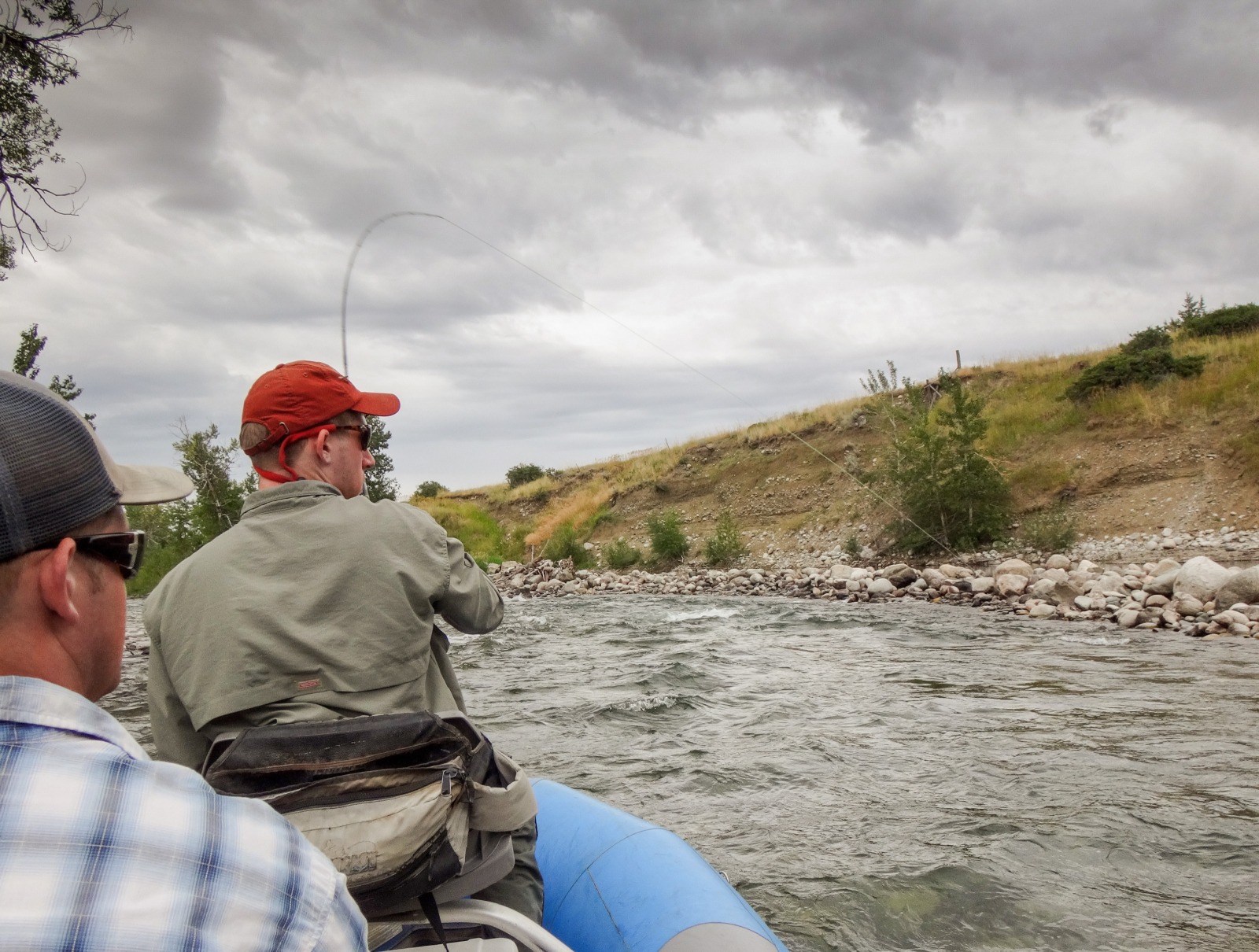 Man in green shirt with a red hat is on a raft with a fishing rod bent because it has hooked a fish, grey cloudy skies in the background with a grassy riverbank.