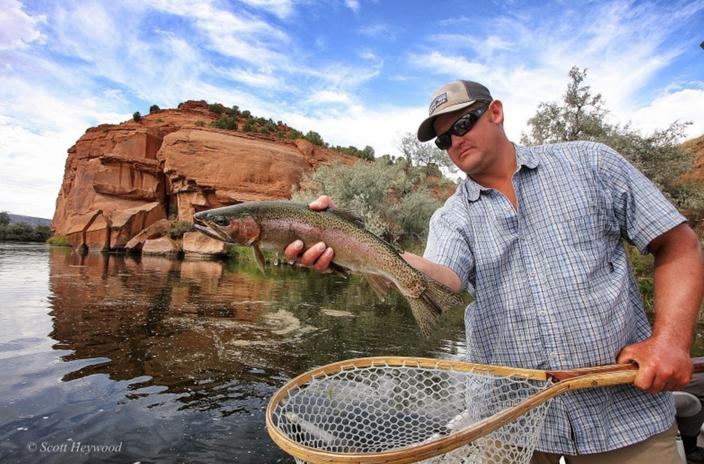 Man holding a rainbow trout above a net with a red clay cliff in the background